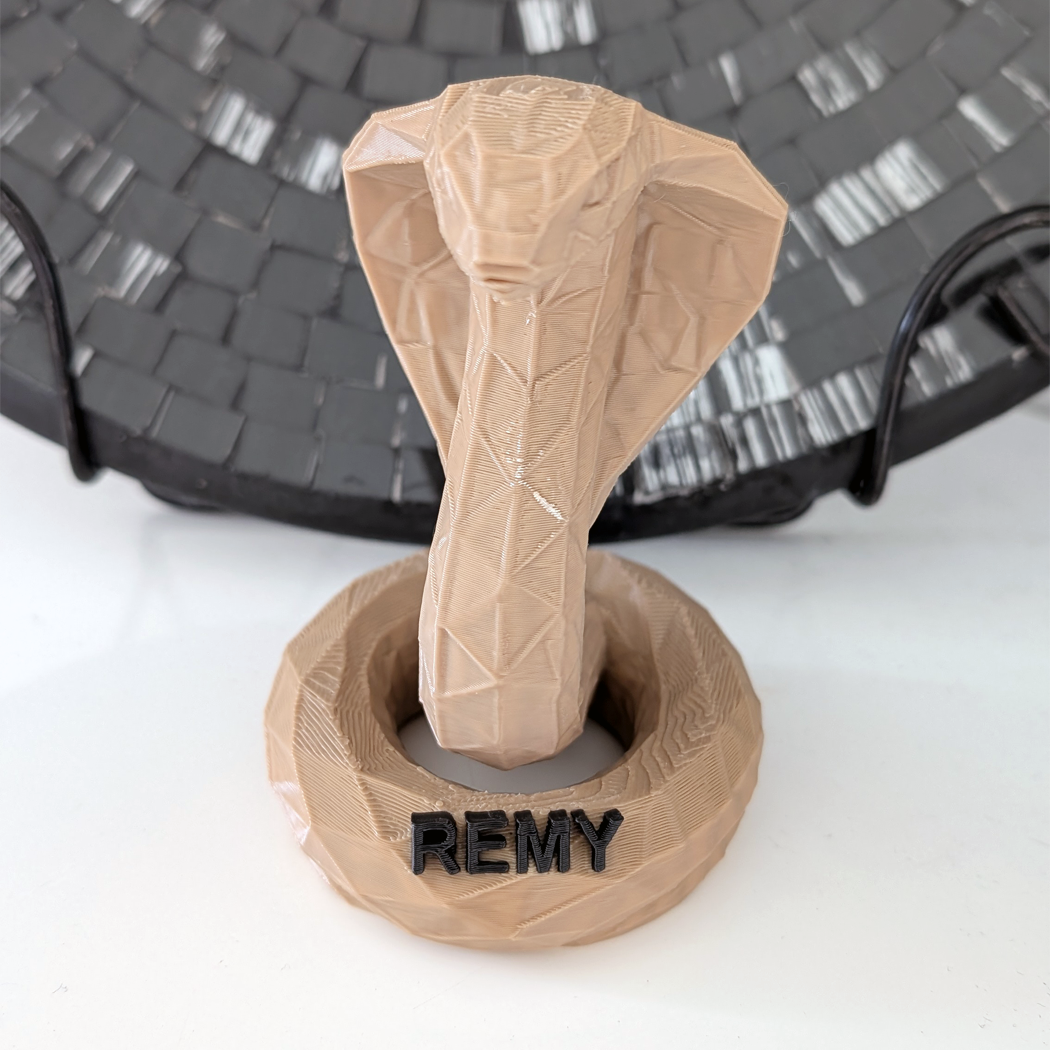 A 3D-printed cobra statue in a light tan color, mounted on a matching circular base with the name 'REMY' printed in black. The statue is displayed on a white surface with a dark woven basket in the background.
