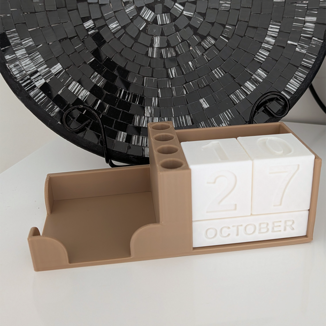 A beige desk organizer calendar with white block numbers showing 27 and October, placed on a white surface with a black circular decorative object in the background.