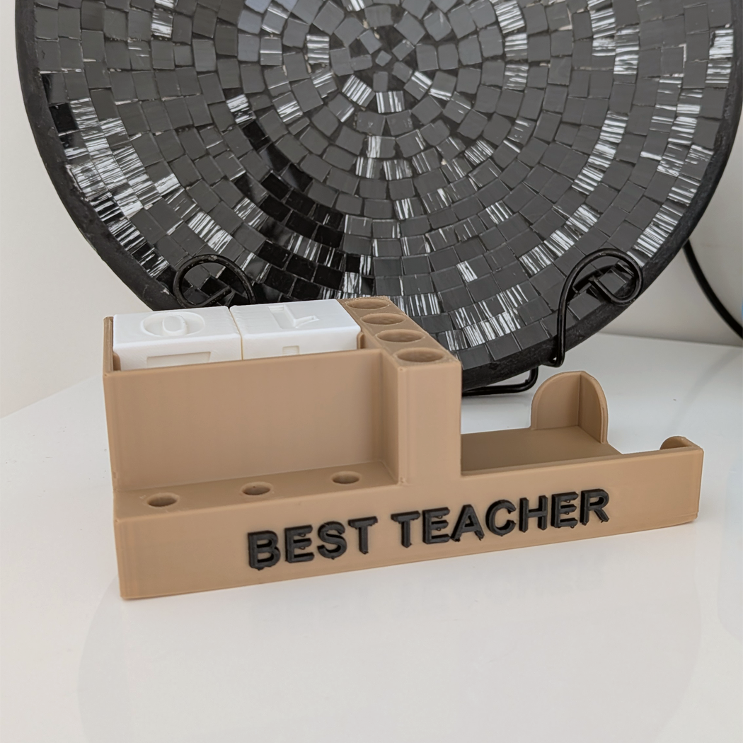 A beige 3D-printed desk sign with the words 'BEST TEACHER' in black, featuring a small white dual-device slot on top, displayed on a white surface with a black mosaic decorative plate in the background.
