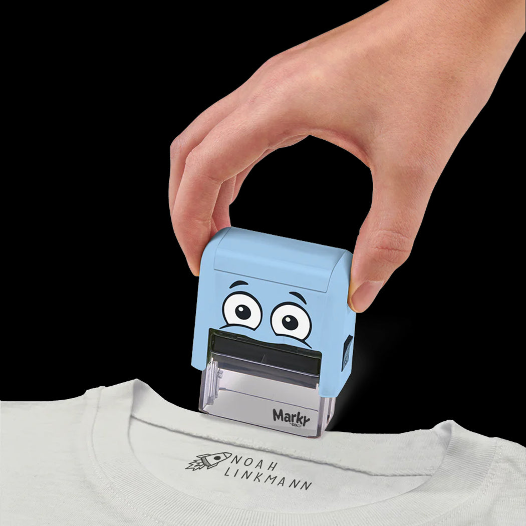 Hand using a blue cartoon-style stamp on a white shirt with 'Noah Linkmann' branding.