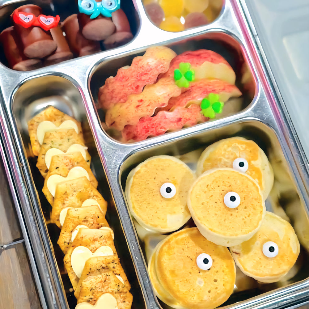 Metal bento box with compartments containing various food items including cookies and small round cakes with eyes.