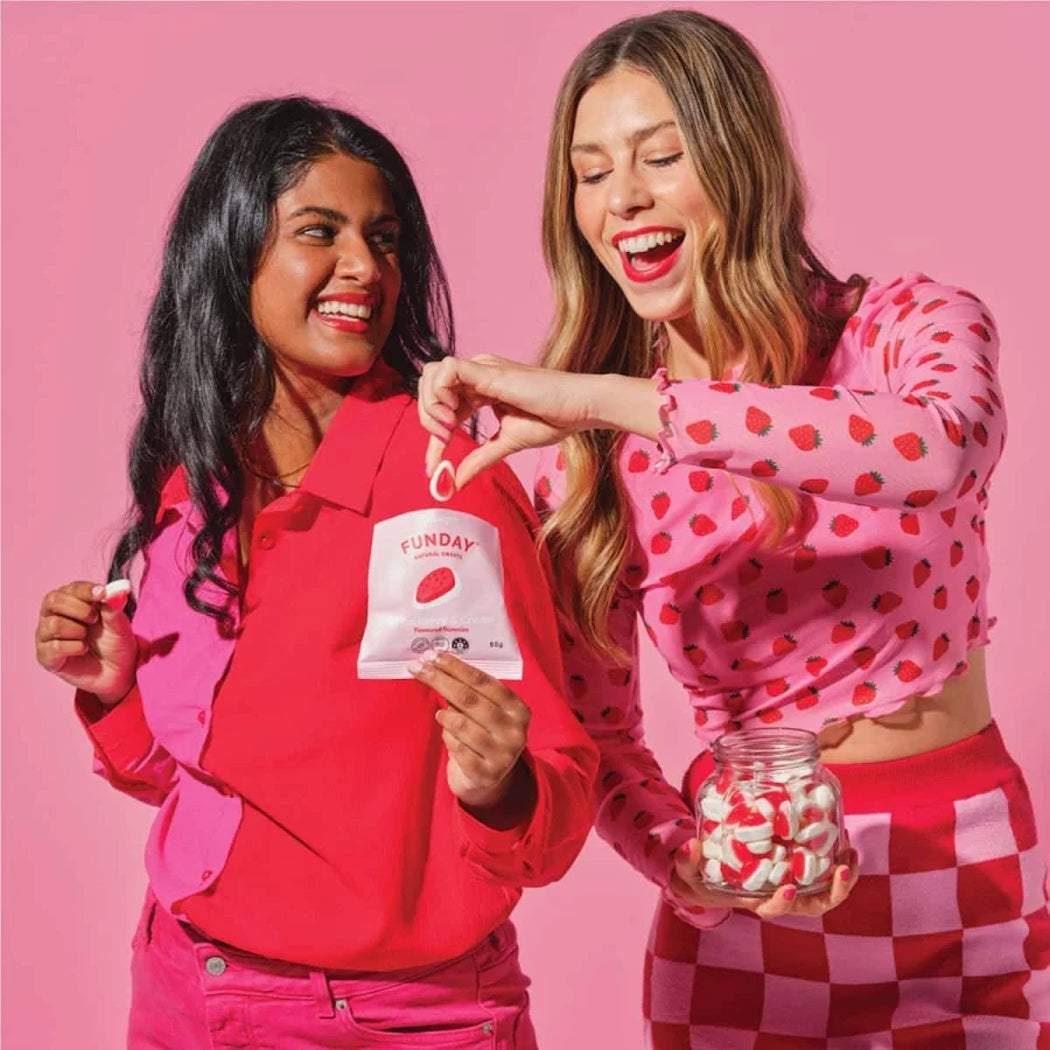 Two women in pink outfits with a pink background, one holding a product and the other a jar of candies.