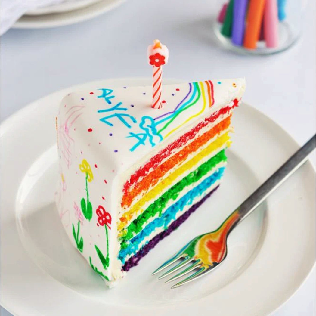 Slice of rainbow cake with a lit candle on a white plate