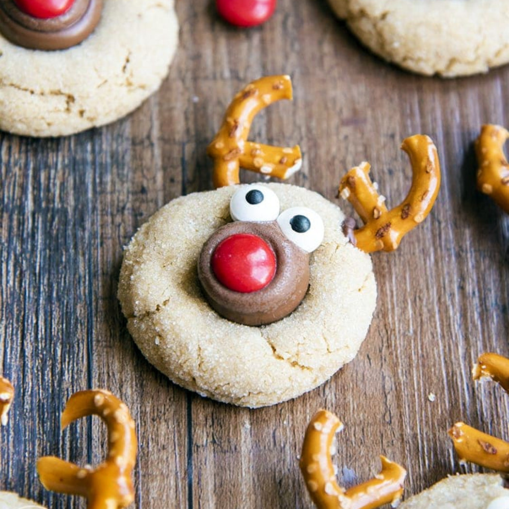 Cookie decorated like a reindeer with pretzel antlers and M&M nose on a wooden surface.