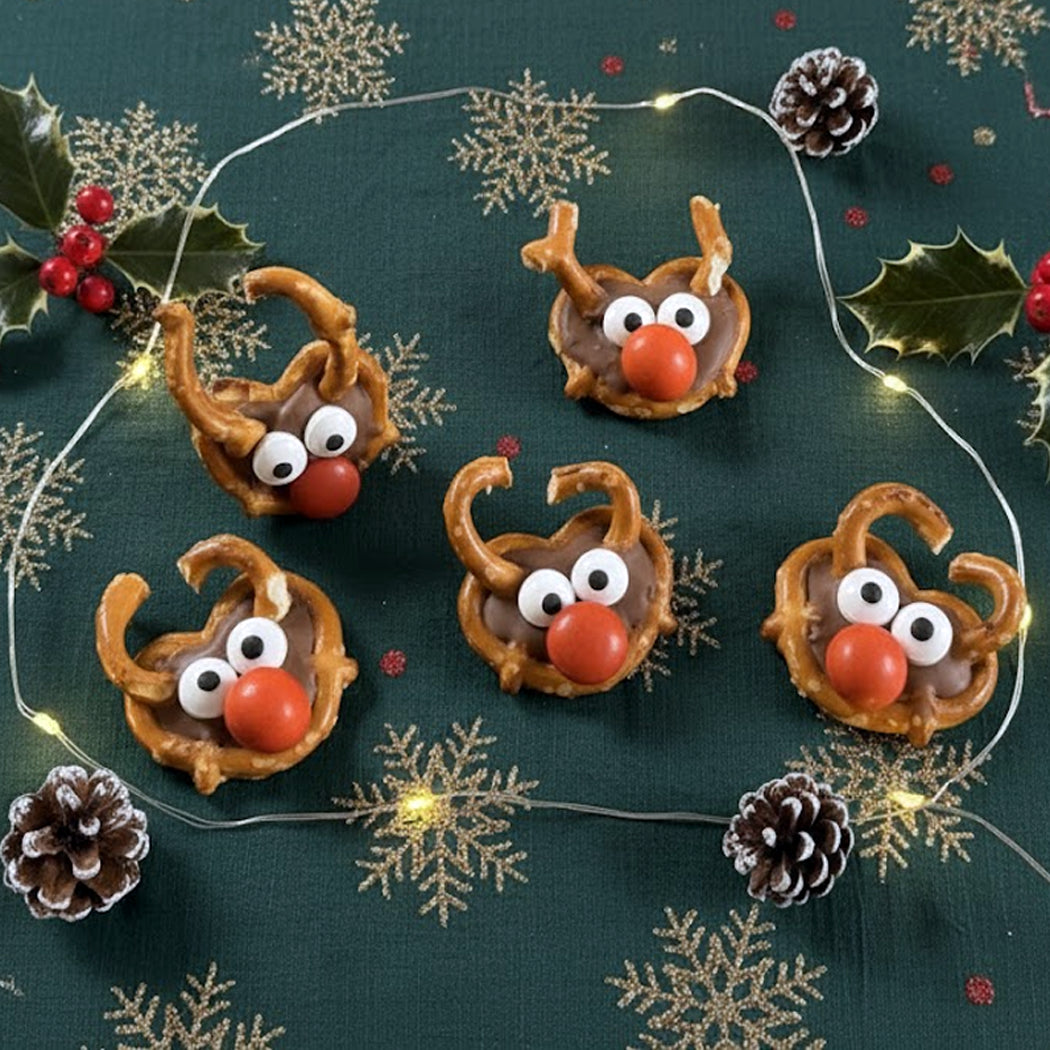 Decorative reindeer pretzels with candy eyes and noses on a festive background with lights and holly.