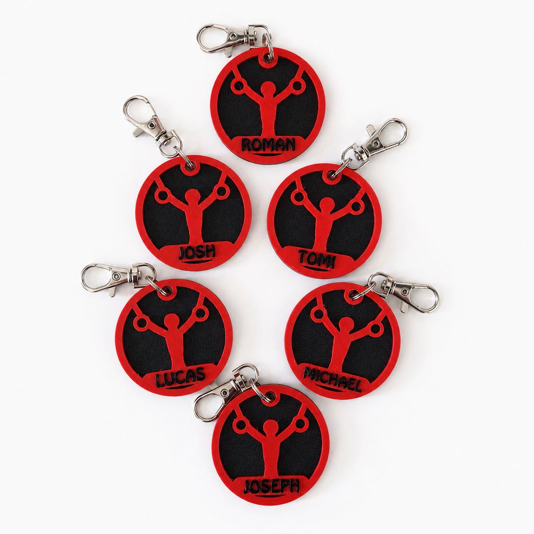 Six personalized round keychains with red and black circular designs featuring a weightlifter silhouette and names (Roman, Josh, Tomi, Lucas, Michael, Joseph), arranged on a white background.