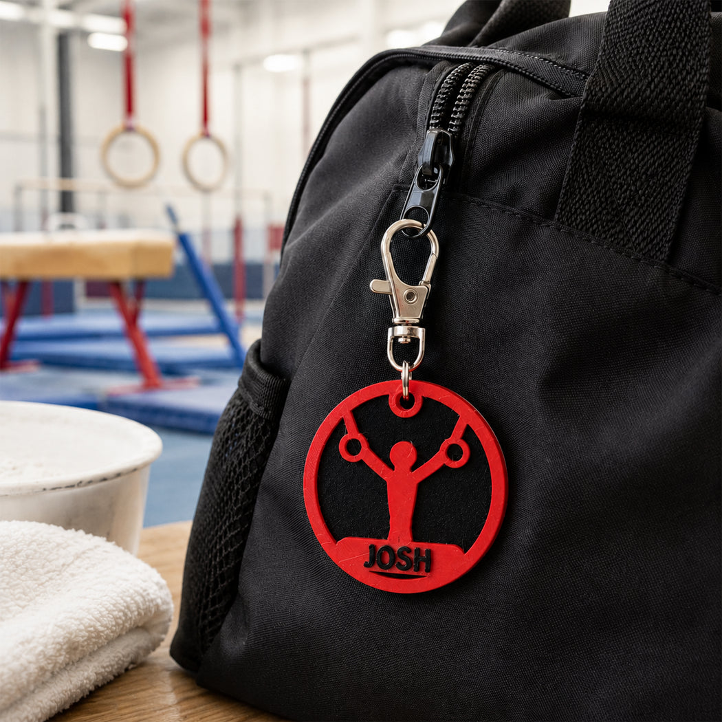 A black gym bag with a red circular custom logo charm featuring a gymnast silhouette and the name 'JOSH' in black, hanging from a silver clasp. The bag is shown on a table in a gym setting with rings and balance beams blurred in the background.