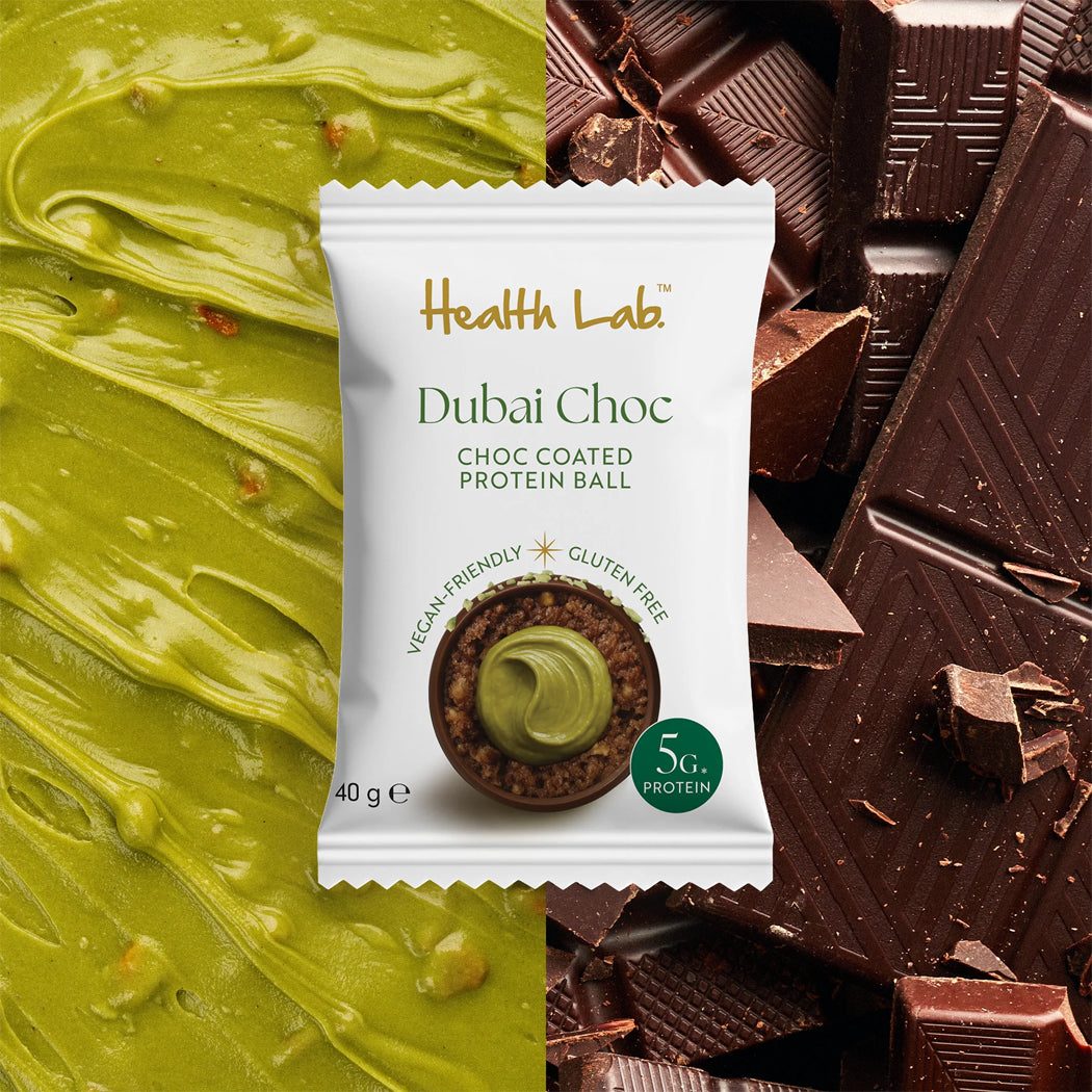 A single packet of Health Lab Dubai Choc Choc Coated Protein Ball centered over a split background: bright green pistachio-like spread on the left and dark chocolate chunks on the right. The packet reads 'Health Lab', 'Dubai Choc', and 'Choc Coated Protein Ball', with a small green '5g protein' badge.