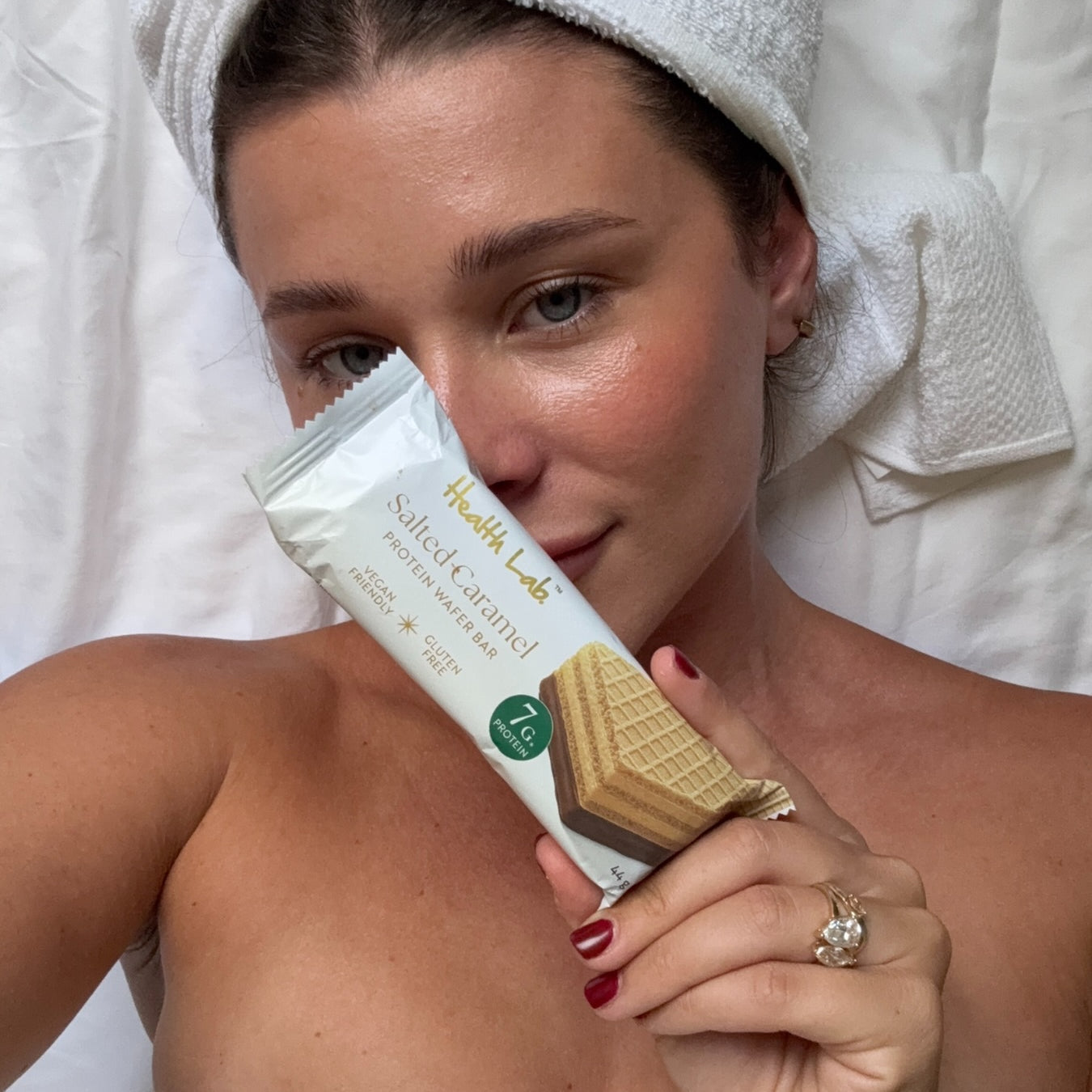 Woman holding a Health Lab salted caramel protein bar with a towel on her head.