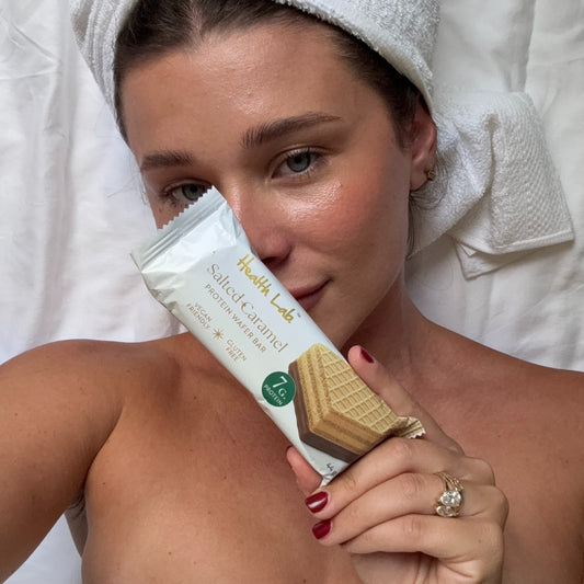 Woman holding a Health Lab salted caramel protein bar with a towel on her head.