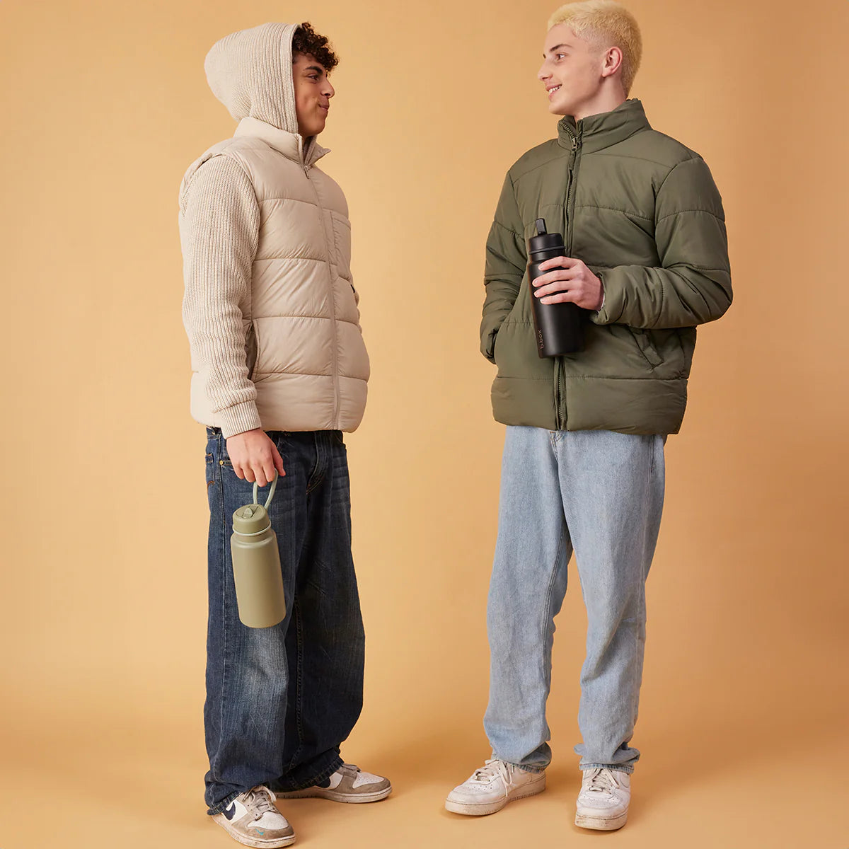 Two people wearing puffer jackets standing against a beige background