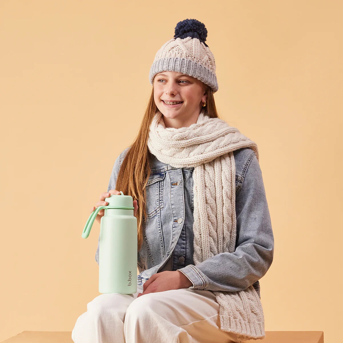 Person wearing a knit hat and scarf, holding a green water bottle against a beige background