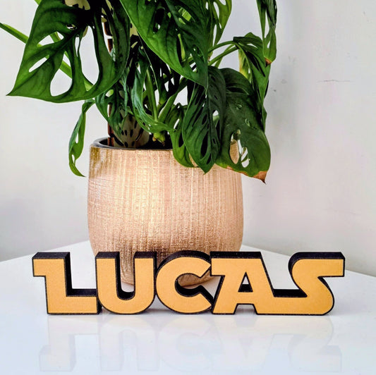 Personalized wooden name sign spelling 'LUCAS' in a warm yellow-gold tone with black 3D lettering, placed on a white surface in front of a potted green plant.