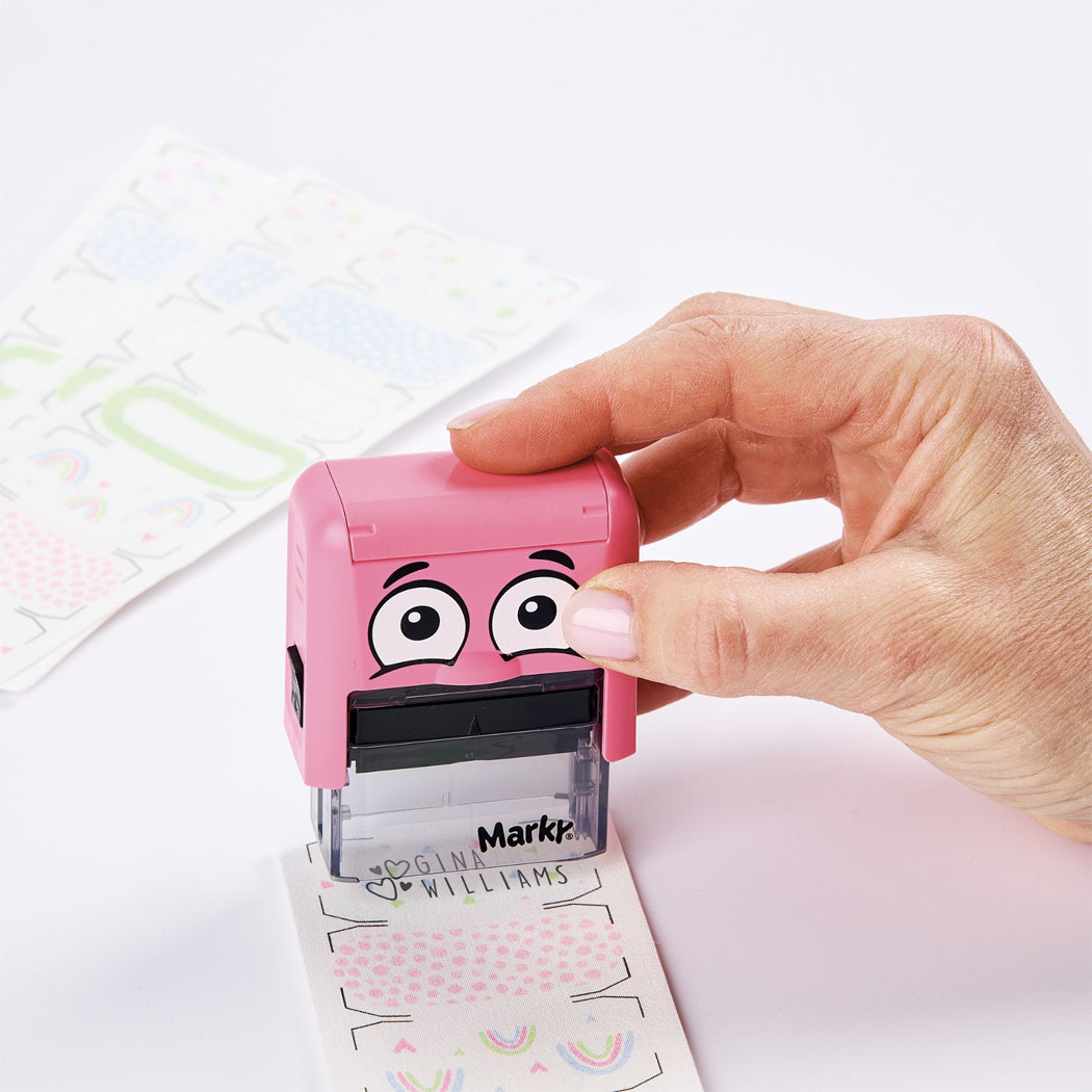 A hand is holding a pink personal stamp with cartoon eyes and the brand name 'Mark' visible on the clear base. The stamp is pressing a sheet of personalized stickers with a pink polka dot pattern and rainbow designs on a clean white background.