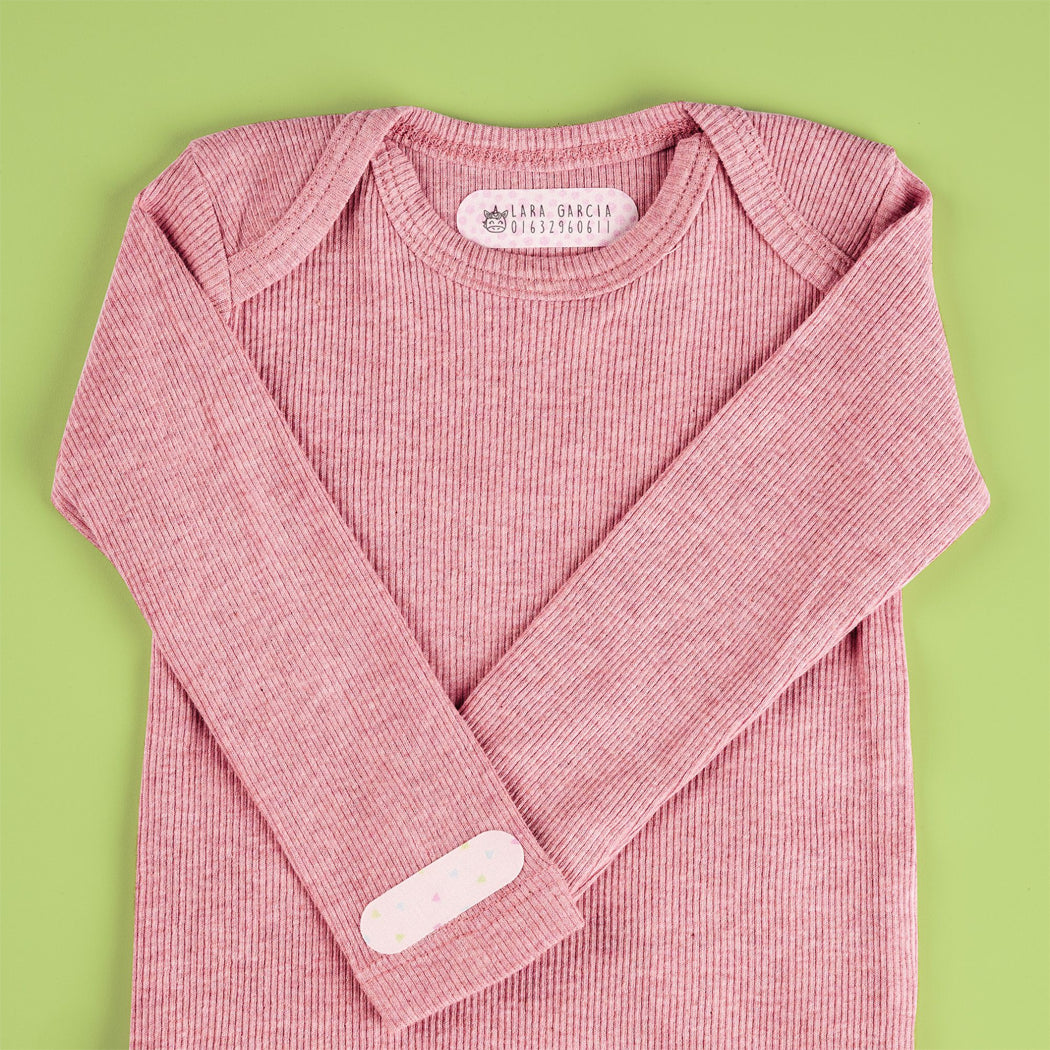 A pink ribbed long-sleeve baby top laid flat on a light green background, with a visible neck label reading 'The Real Grace Baby Clothes' and a small white patch on one sleeve.