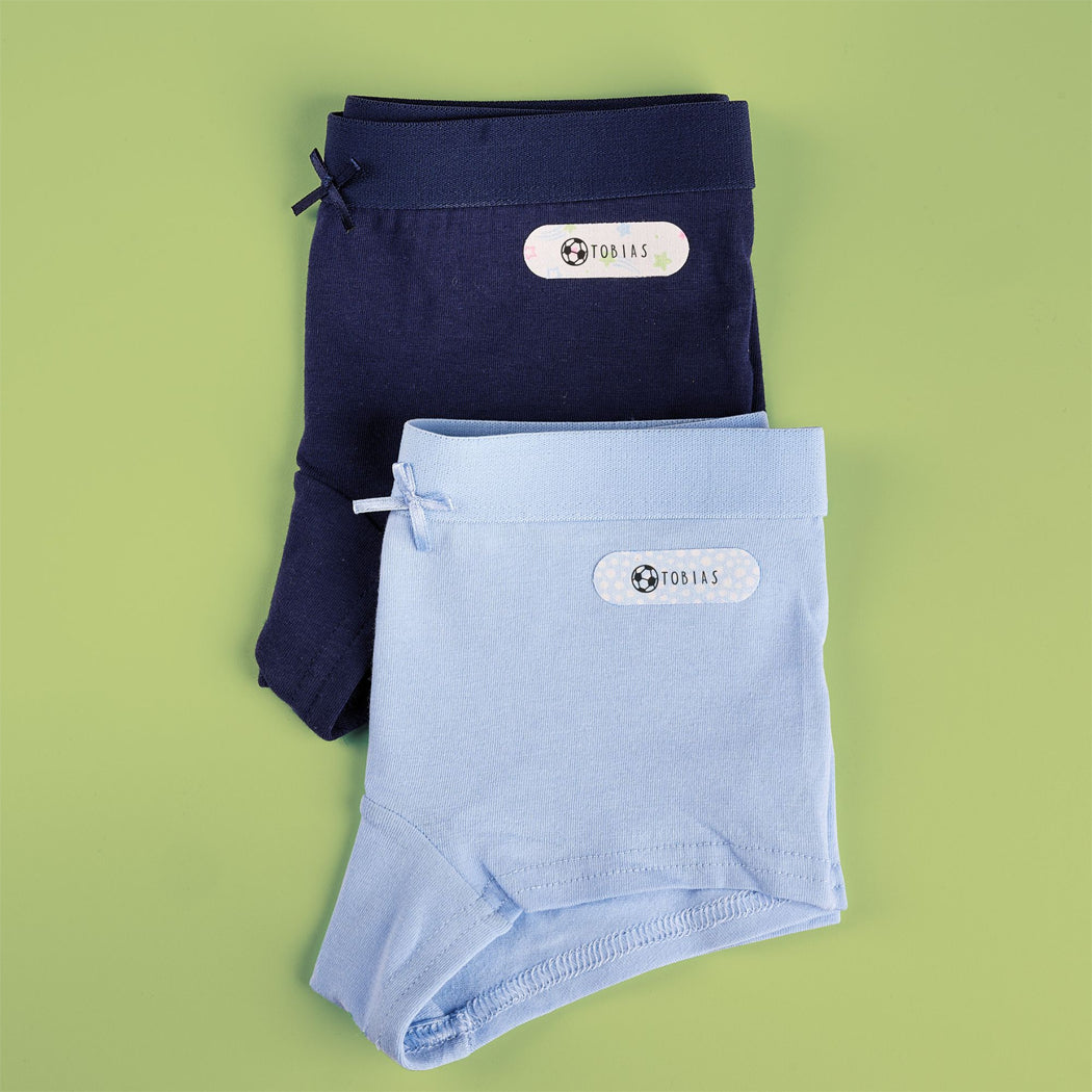 Two pairs of folded children's shorts in navy blue and light blue, arranged on a solid light green background. Each pair has a white oval label with the brand name 'TOP143' and a small soccer ball logo.