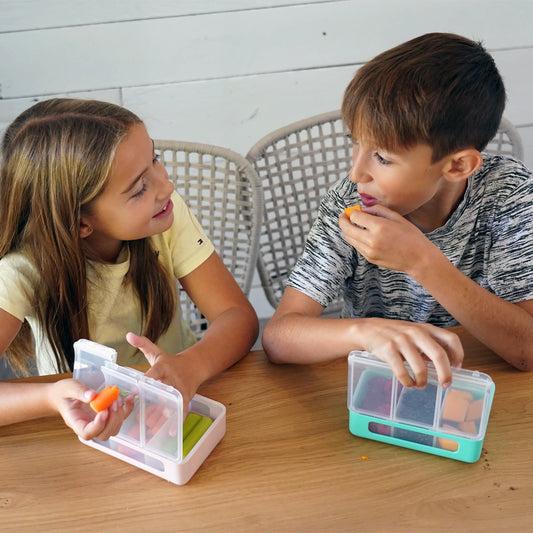Two children at a table with lunch boxes, one eating a carrot.