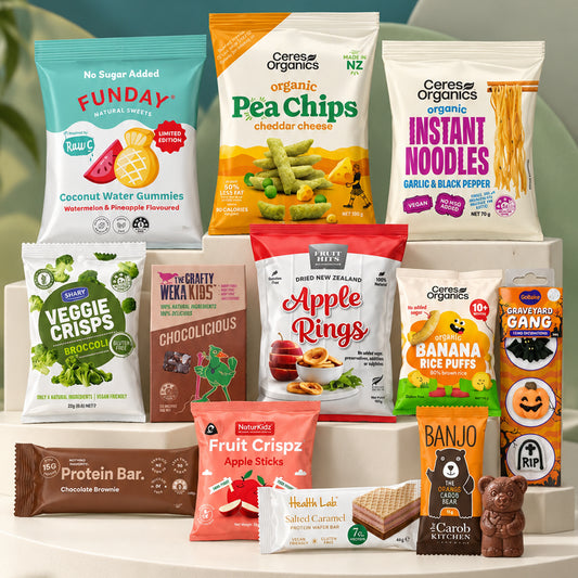 A grouped assortment of packaged snack foods and a protein bar arranged on white display blocks against a light gray studio background. Visible products include Ceres Organics Pea Chips, Ceres Organics Instant Noodles, Funday Coconut Water Gummies, Veggie Crisps, Apple Rings, Banana Rice Puffs, Health Lab Fruit Crispz, Banjo Chocolate, and a Protein Bar.