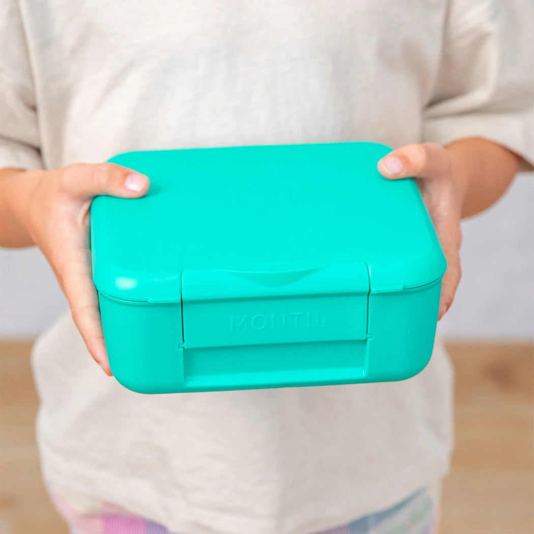 Person holding a teal lunch box with 'Yumbox' branding.