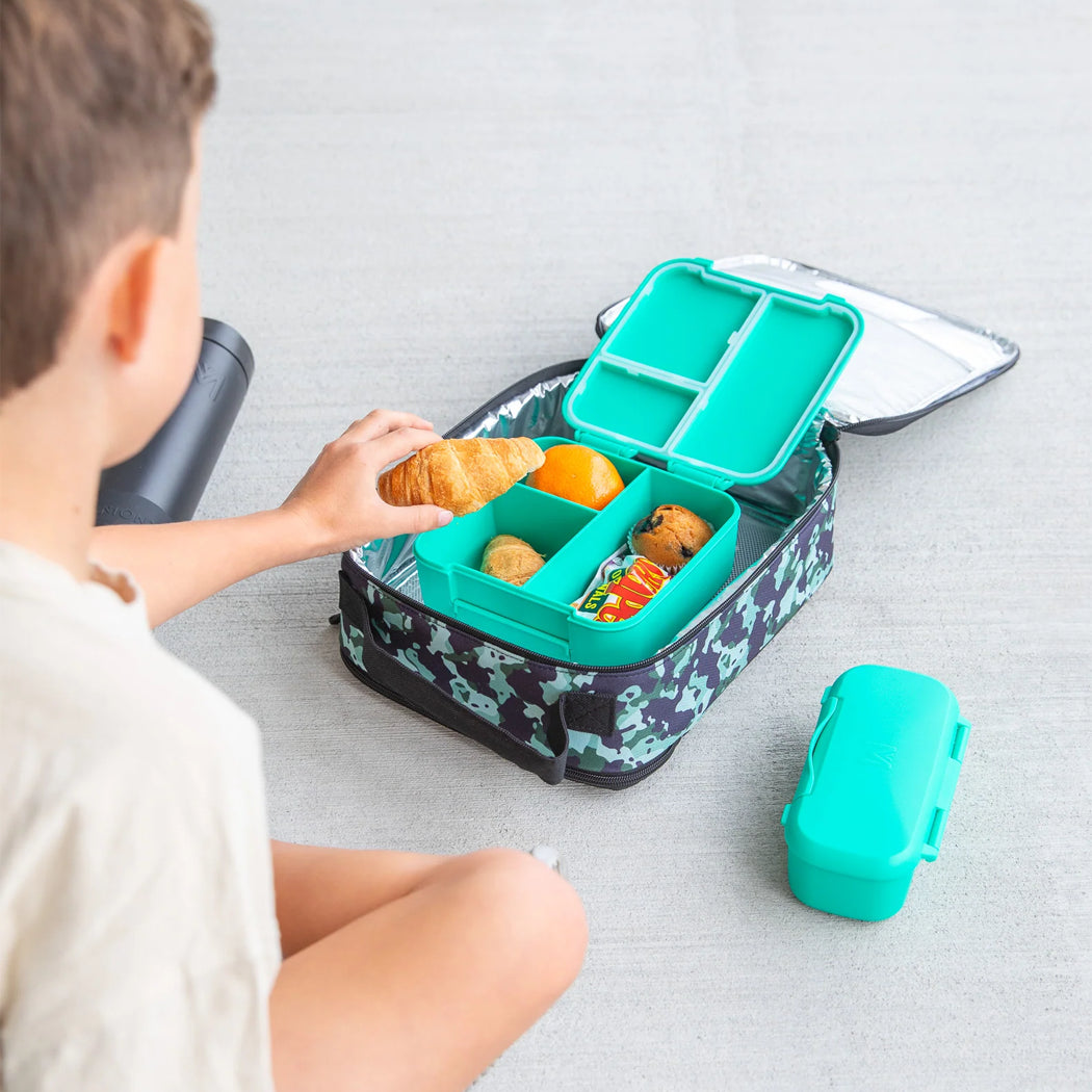 Child reaching into a bento-style lunch box with compartments filled with food on a light gray surface.