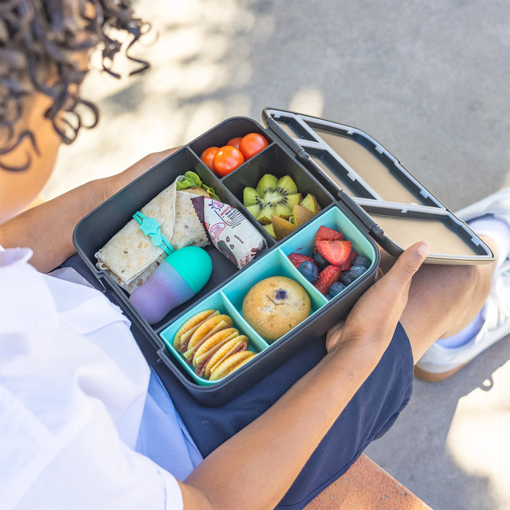 Person holding a bento box with various food items outdoors