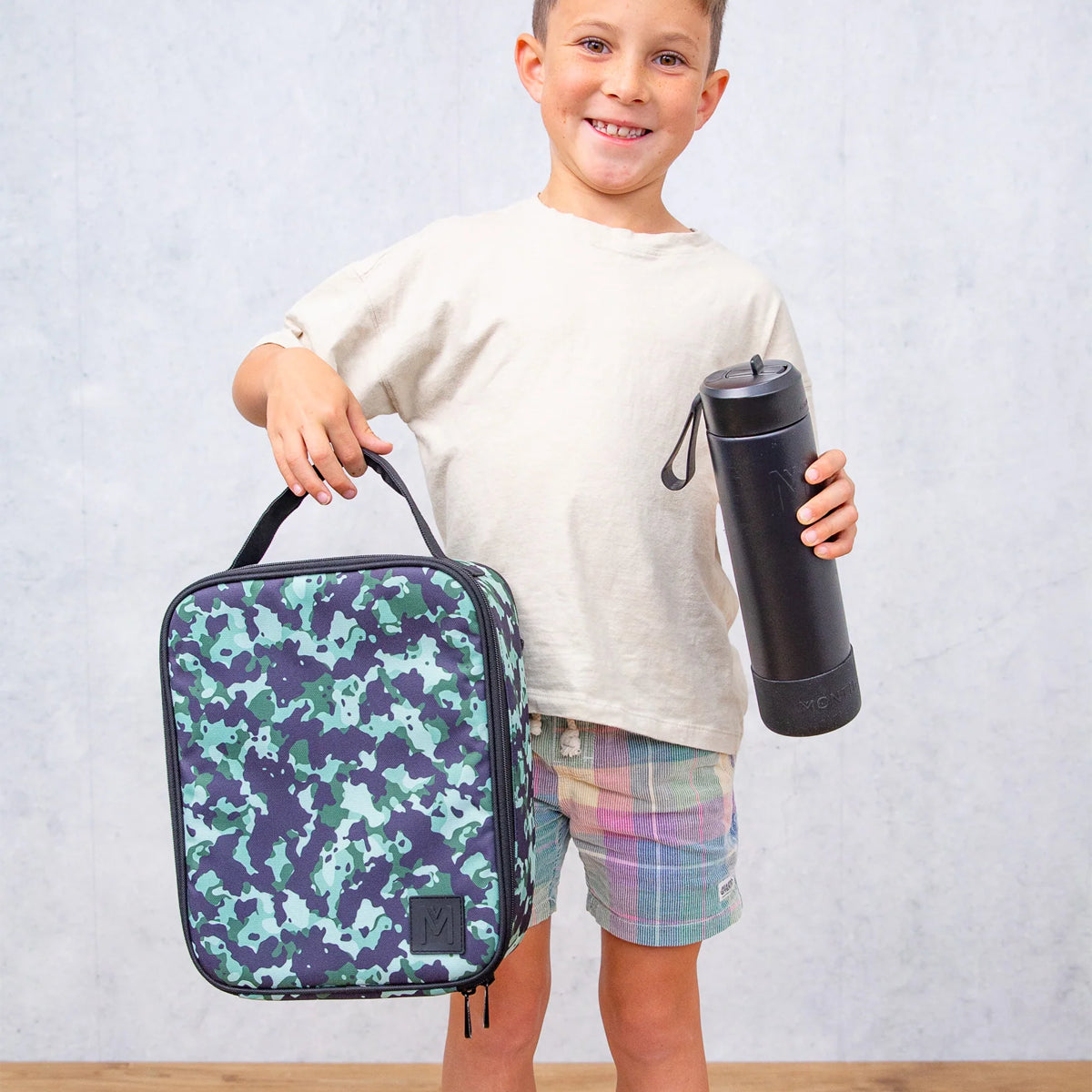 Child holding a camouflage backpack and a black tumbler against a light gray background