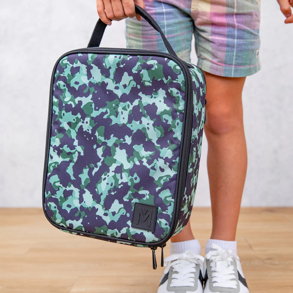 Person holding a green and purple camouflage lunch bag on a wooden floor.