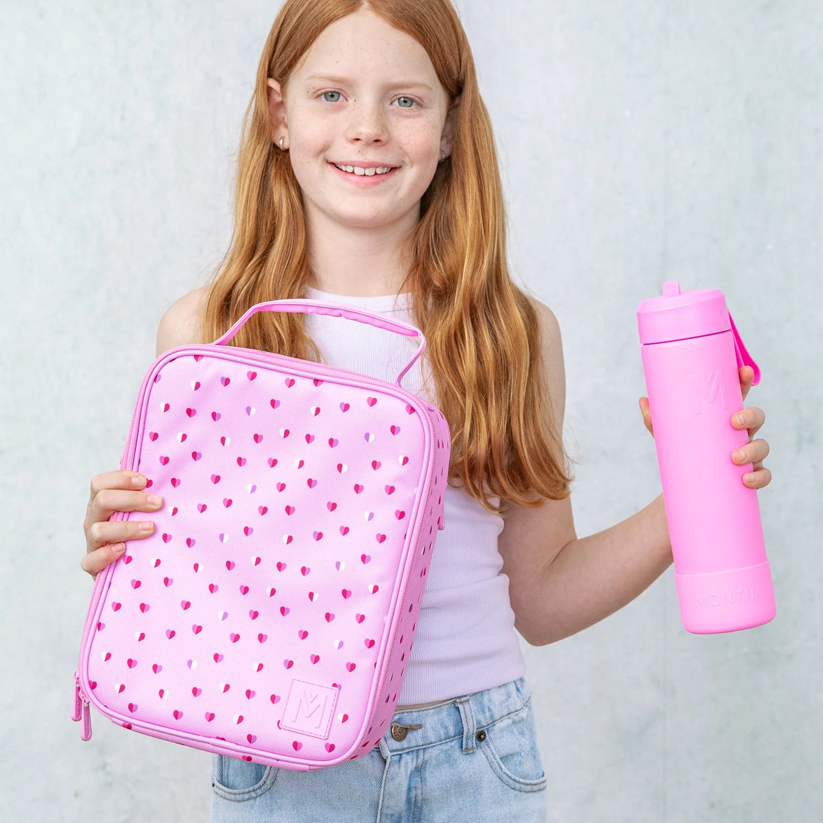 Girl holding a pink heart-patterned lunch bag and a pink water bottle against a light background