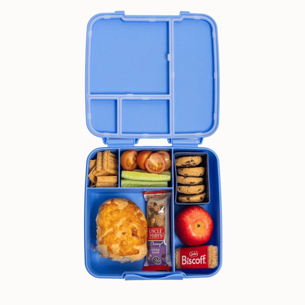 A blue bento lunch box with multiple compartments, shown open and filled with snacks and food items on a plain white background. Visible packaged snacks include Uncle Arthur's and Biscuit.
