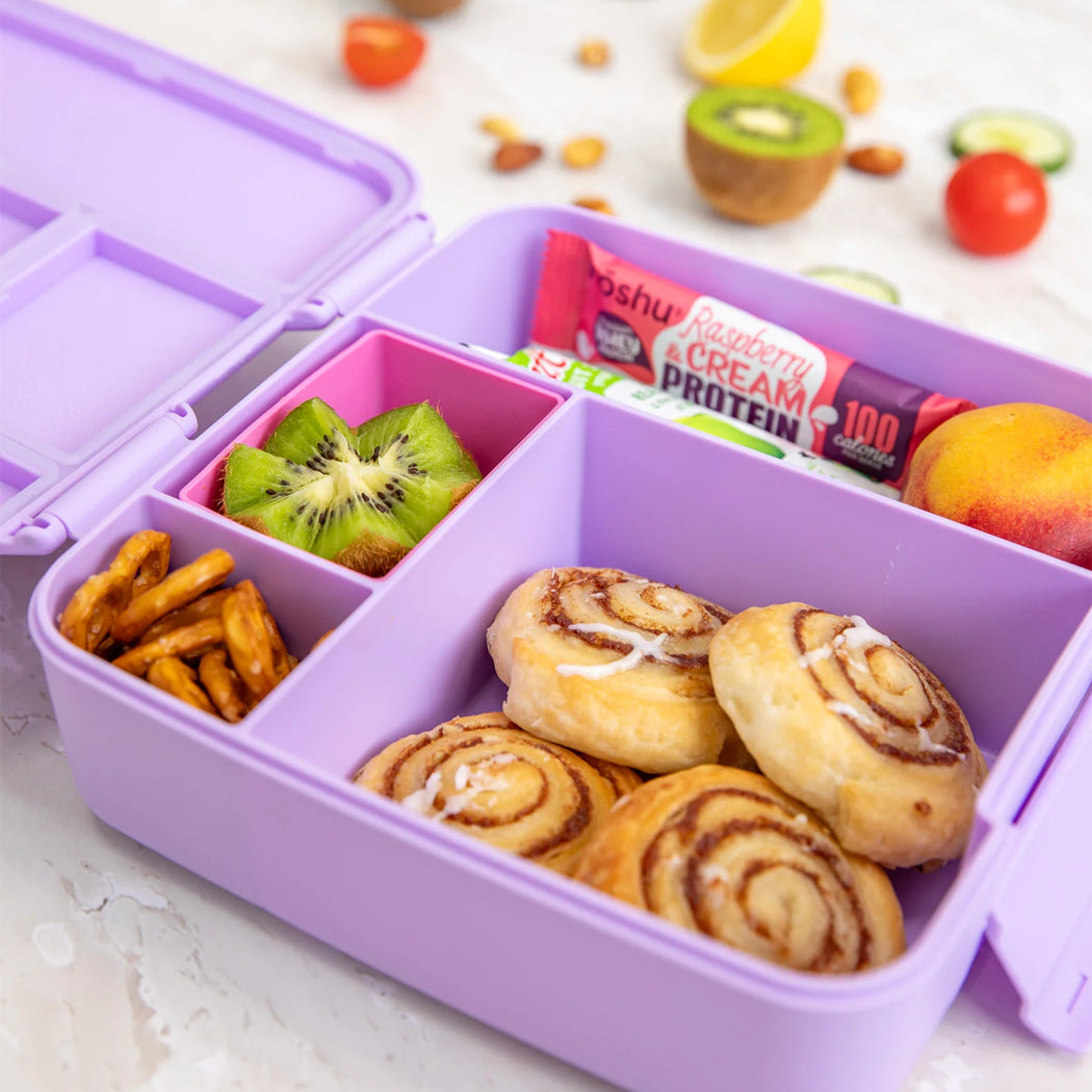 Purple bento box with snacks including kiwi, cookies, and a protein bar on a white surface.