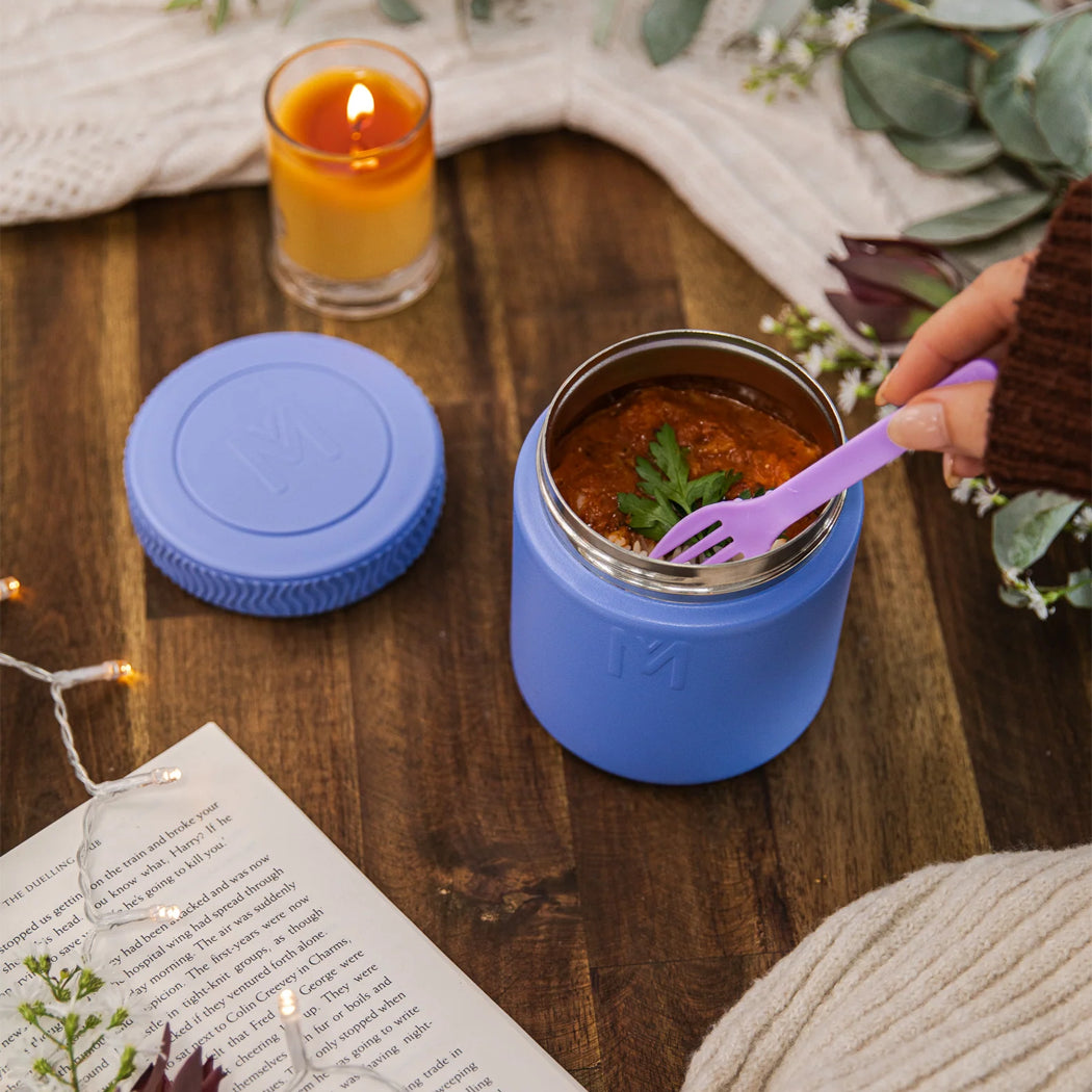 Blue insulated food container with a purple spoon on a wooden table, surrounded by a lit candle and a book.