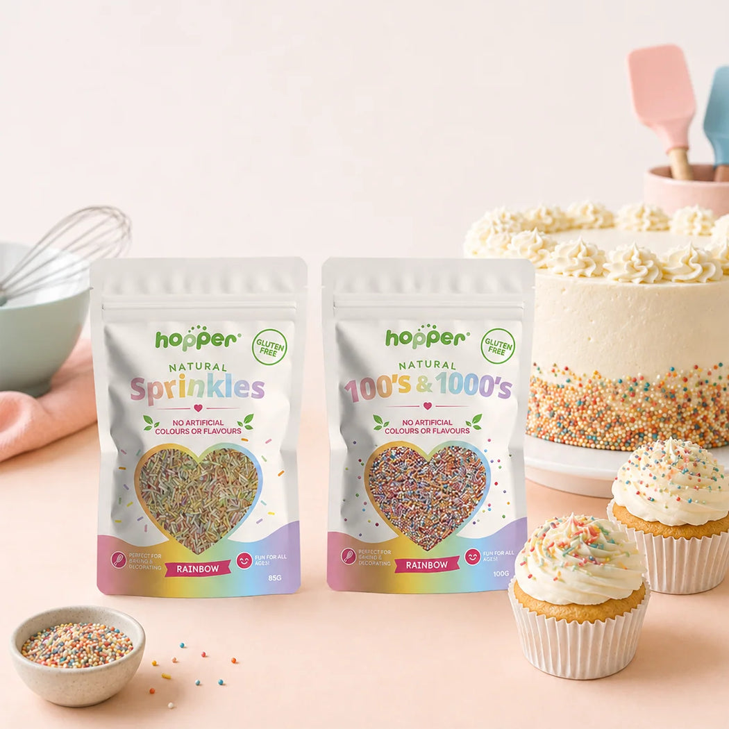 Two bags of hopper Natural Sprinkles displayed on a pink surface next to a decorated cake and cupcakes, with a small bowl of loose sprinkles in the foreground.