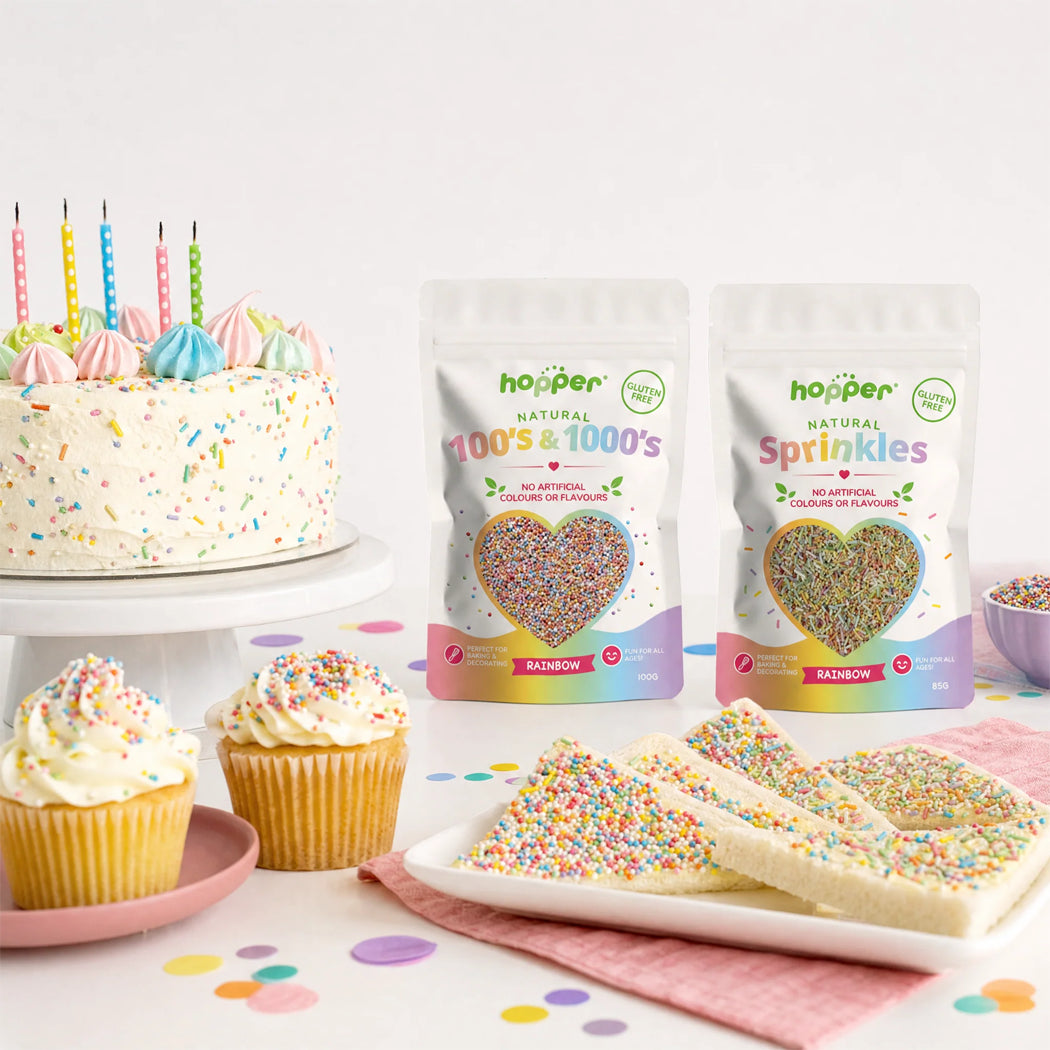 Two bags of hoppen Natural 100's & 1000's Sprinkles displayed on a white table next to decorated cupcakes, a birthday cake with candles, and sprinkle-coated cookies, with confetti scattered on the surface.