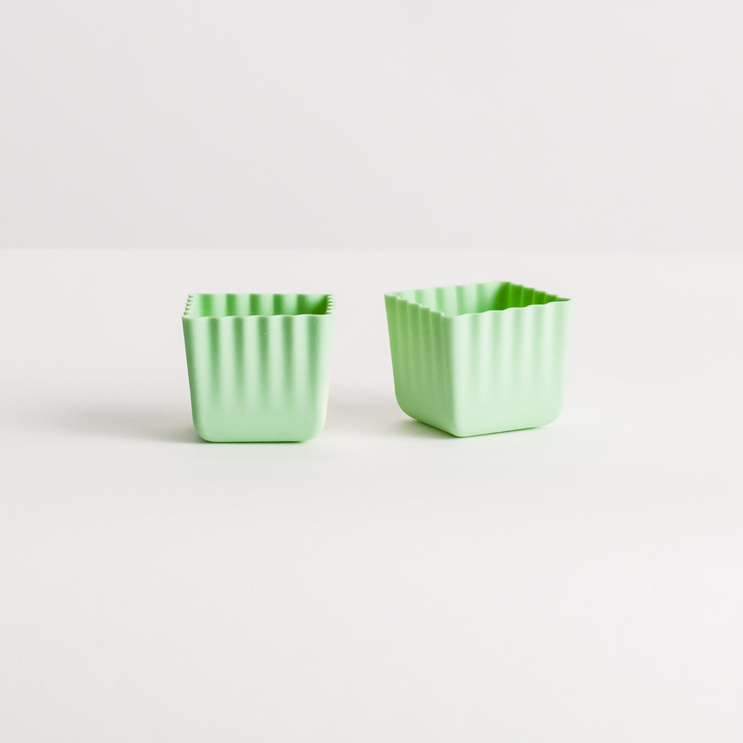 Two small mint green square silicone pots with a vertical ribbed texture, standing side-by-side on a clean white surface against a light gray background.