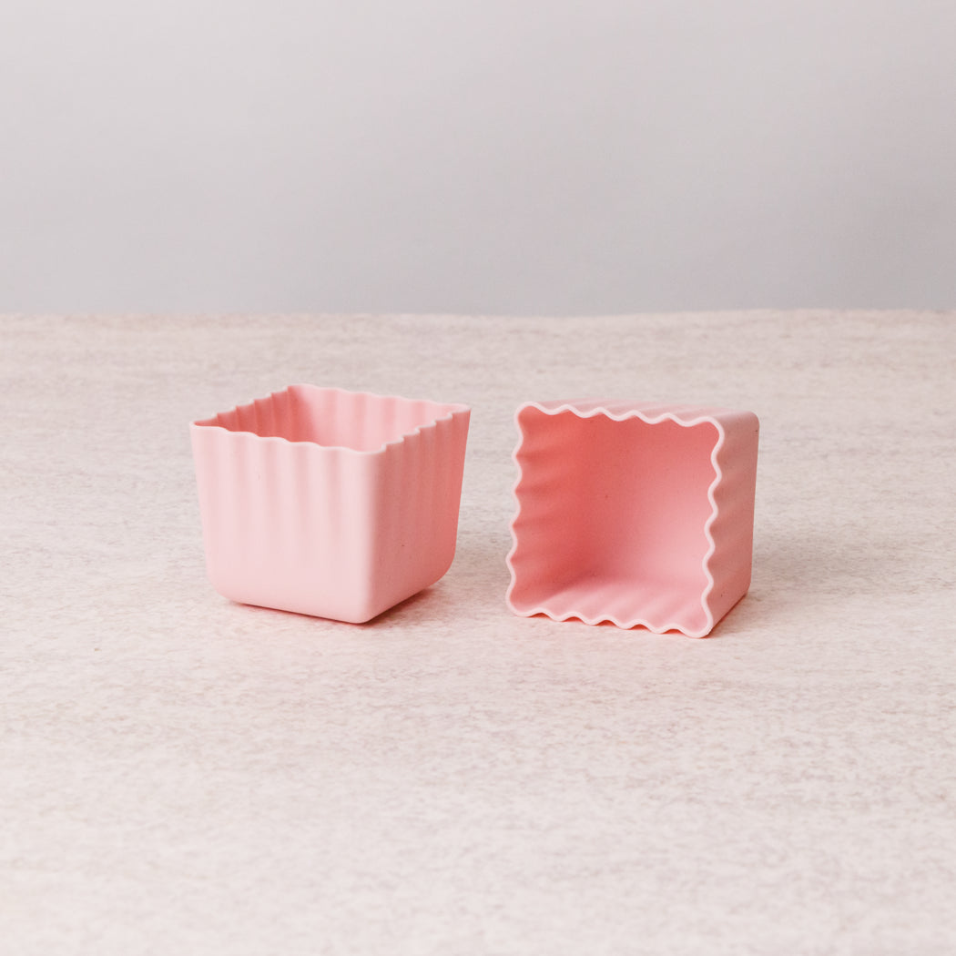 Two small pastel pink square silicone baking molds with wavy scalloped edges, shown on a light beige textured surface against a neutral gray background.