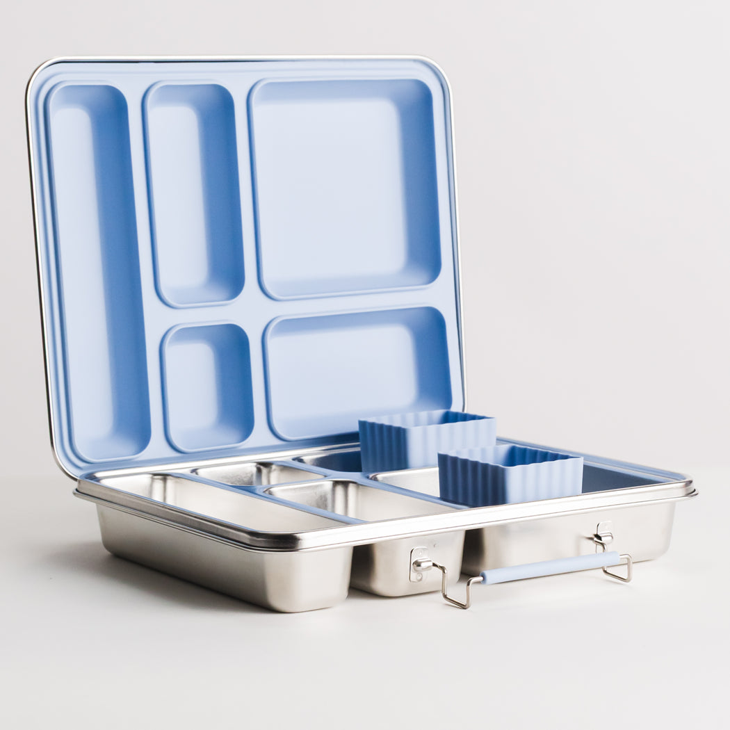 A stainless steel lunch box with an open lid, showing a light blue compartmentalized interior and two small blue silicone cups, photographed on a clean light gray background.
