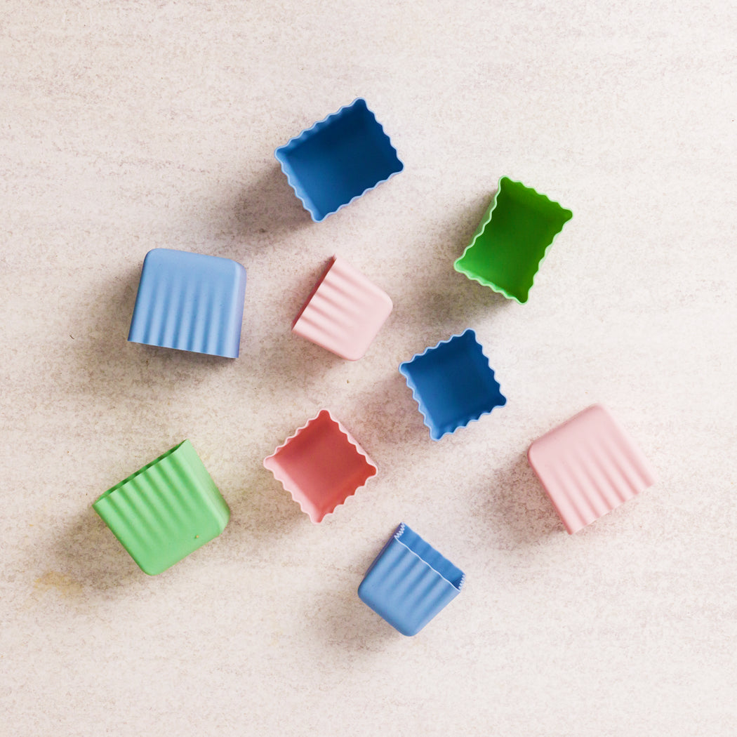 Nine small square silicone baking molds in assorted pastel and bright colors, arranged on a light beige textured surface.
