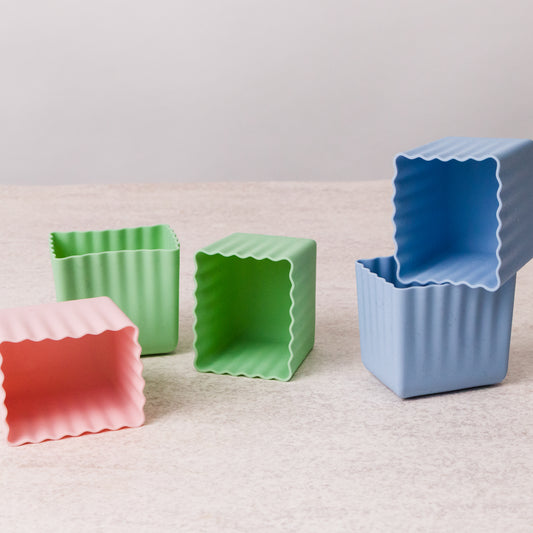 Four square, open-top silicone baking molds with wavy scalloped edges in pink, green, and blue, arranged on a light beige textured surface against a neutral light gray background.