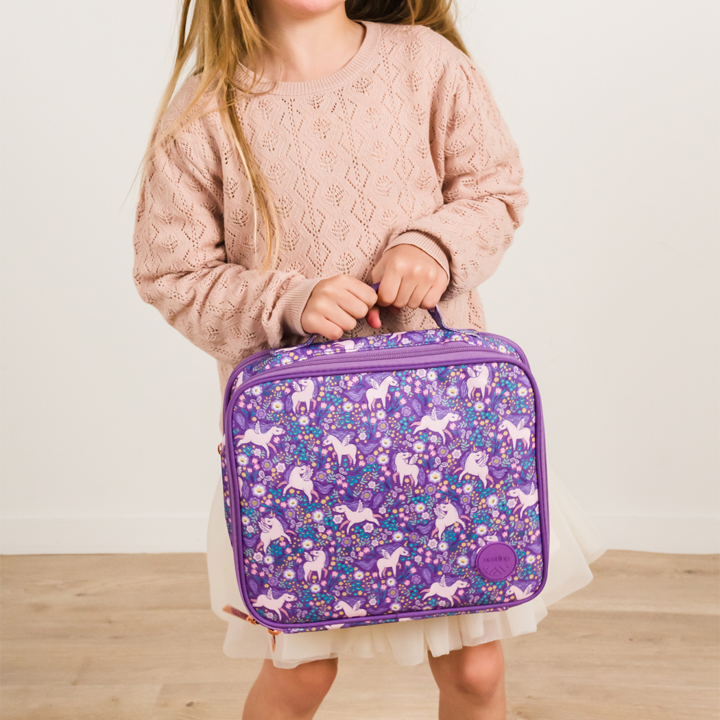 A young girl in a pink sweater holds a purple insulated lunch bag decorated with a white unicorn and floral pattern, set against a neutral indoor background.