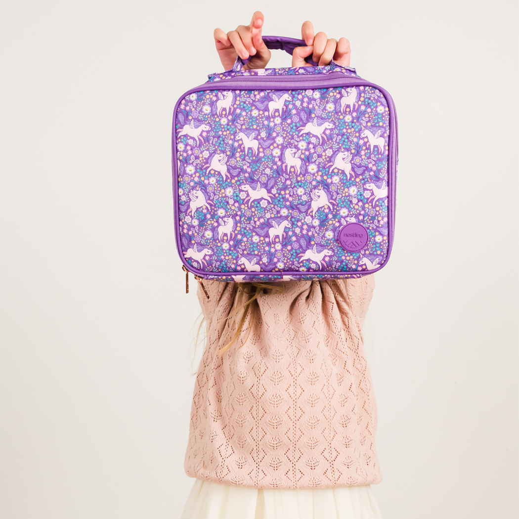 A child holds up a purple lunch bag with a unicorn pattern, showing their hands and the top of their pink textured sweater against a plain light background.