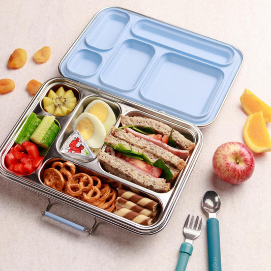 Lunchbox with compartments filled with food, surrounded by fruits and utensils on a light surface.