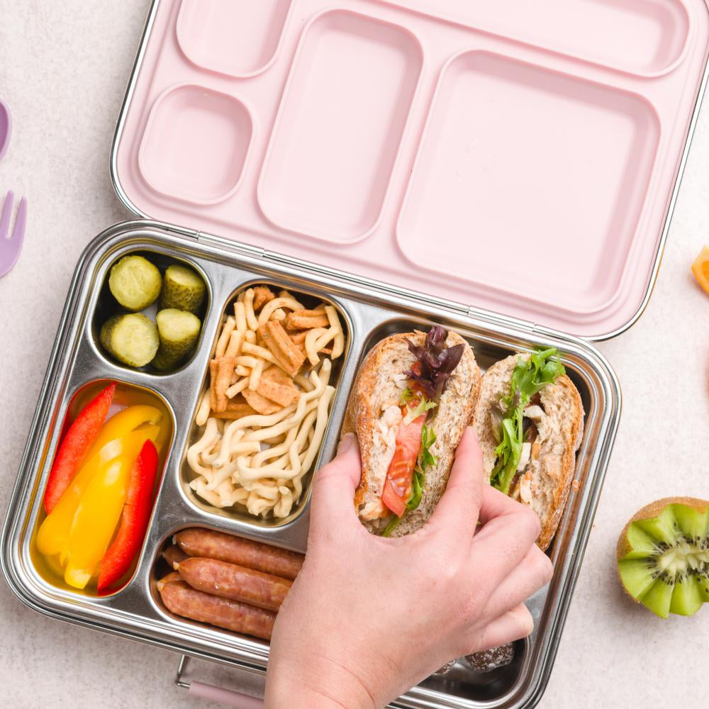 Bento box with compartments containing a sandwich, noodles, pickles, and vegetables on a light surface.