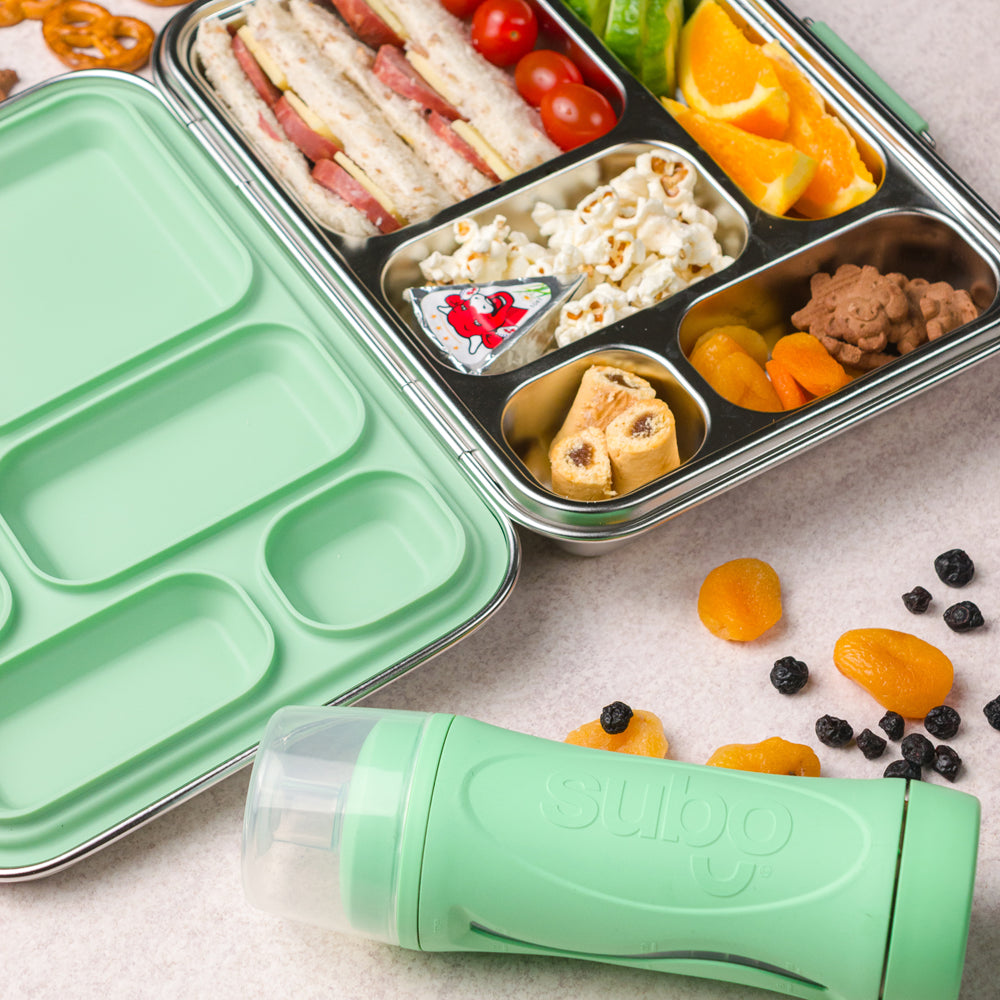 Lunchbox with compartments filled with food and a green container labeled 'Subo' on a light surface.