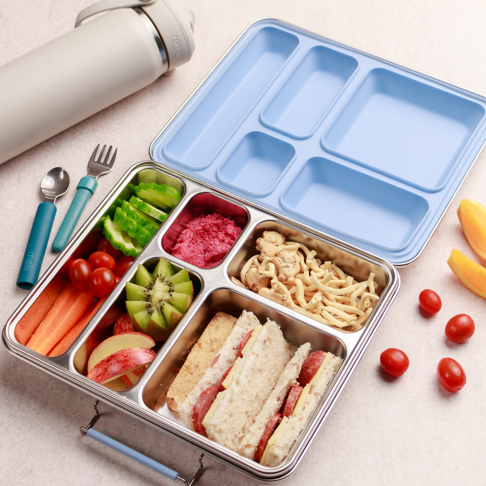 Divided lunch container with food compartments, including sandwiches, fruits, and vegetables, on a light surface.