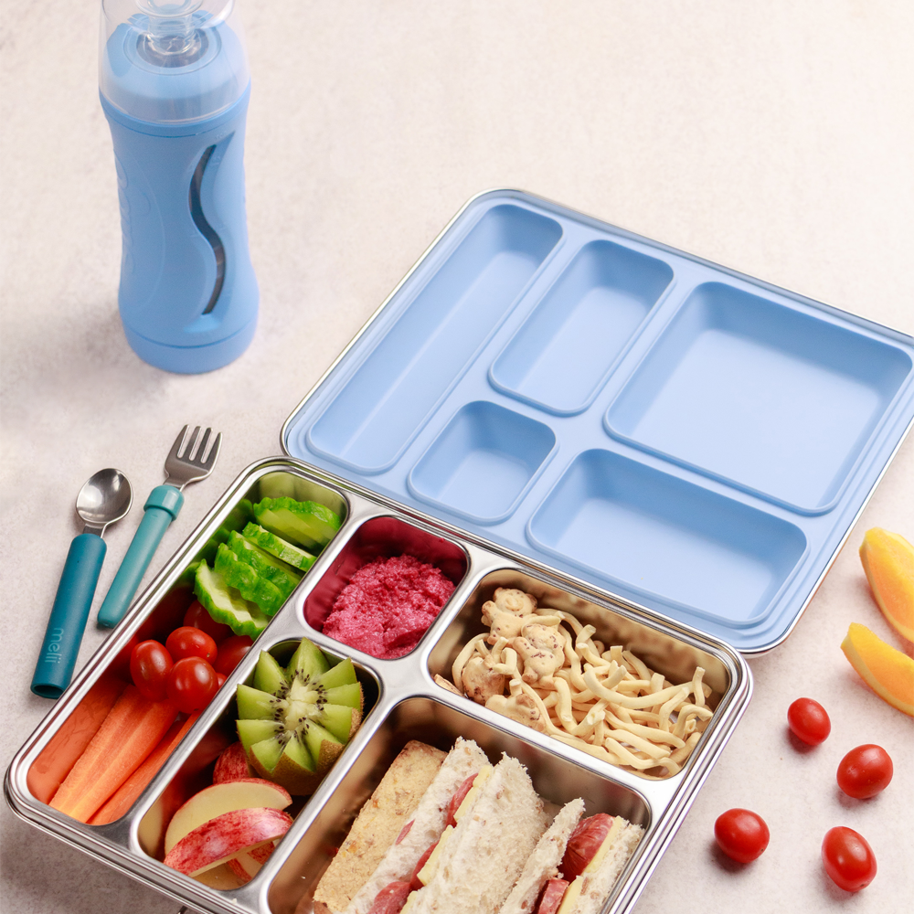 Divided lunch container with compartments filled with food, next to a blue water bottle and utensils on a light surface.