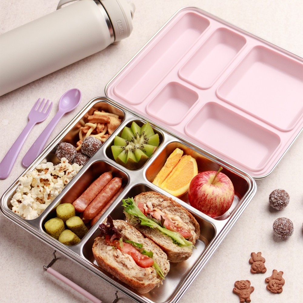 Divided lunch container with food items including a sandwich, fruit, and snacks, next to a water bottle and utensils on a light surface.