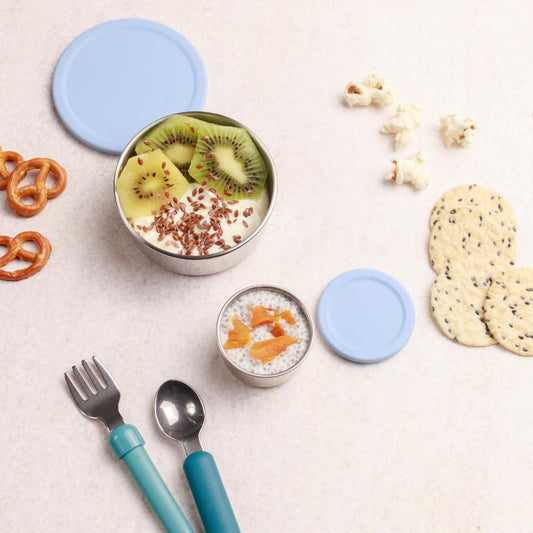Two small containers with lids, a bowl of fruit, and snacks on a light surface.