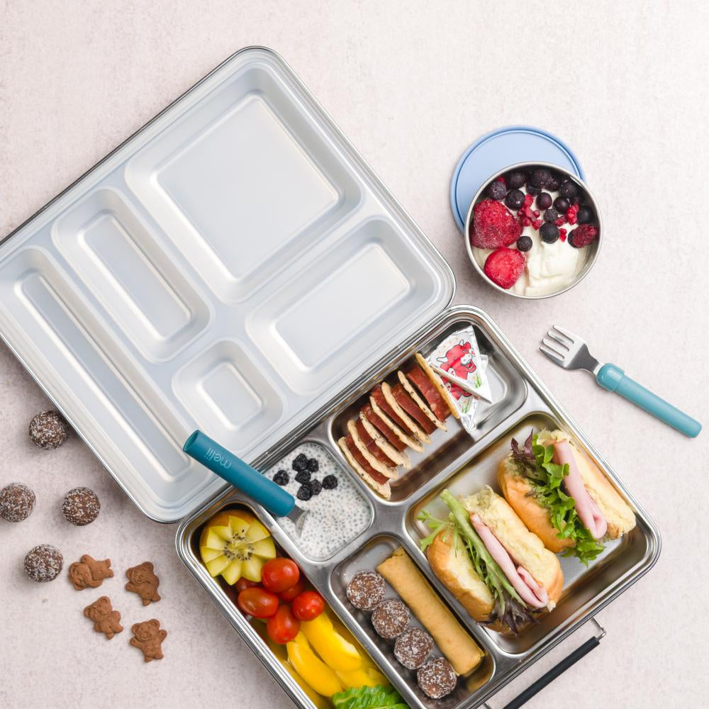 Metal lunch box with compartments filled with food on a light surface.