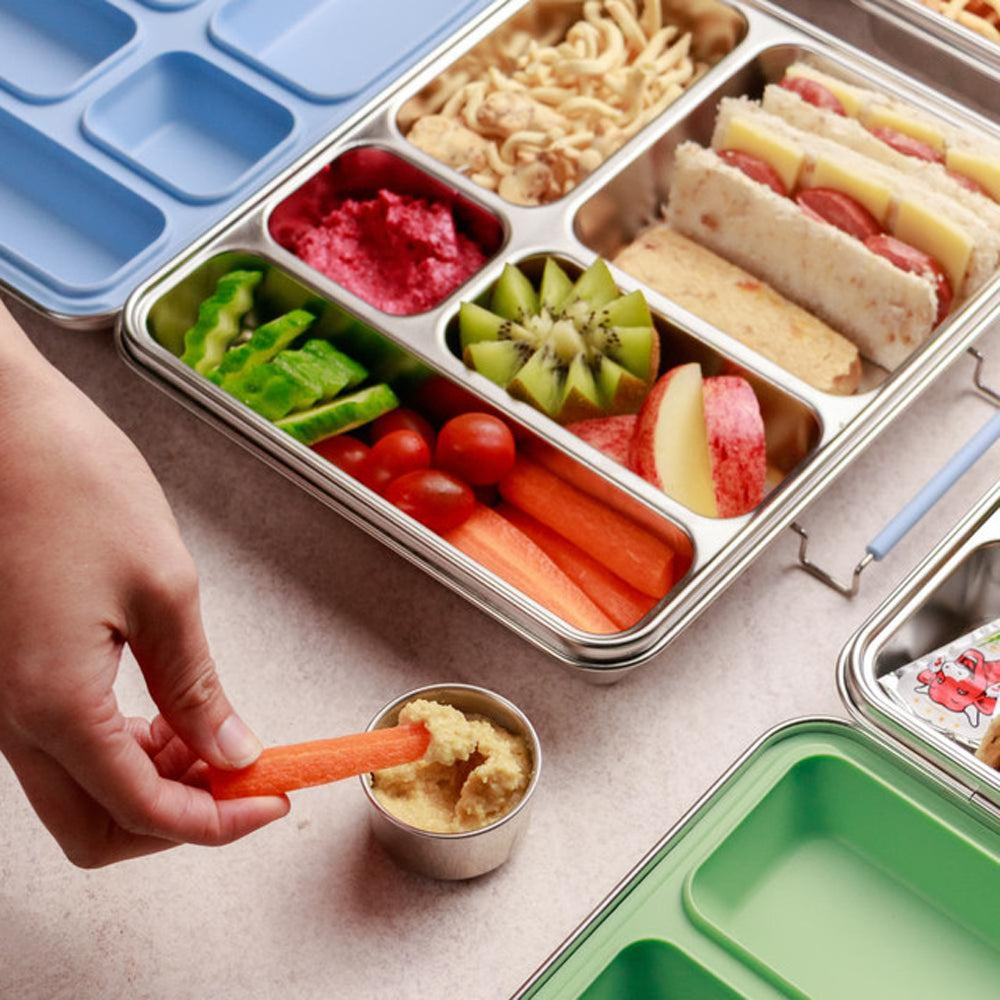 Metal lunch box with compartments filled with various food items and a hand dipping a carrot stick into a small container.