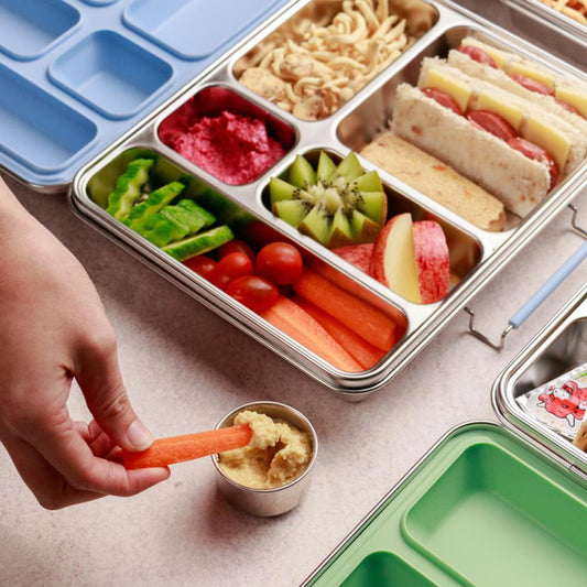 Metal lunch box with compartments filled with various food items and a hand dipping a carrot stick into a small container.